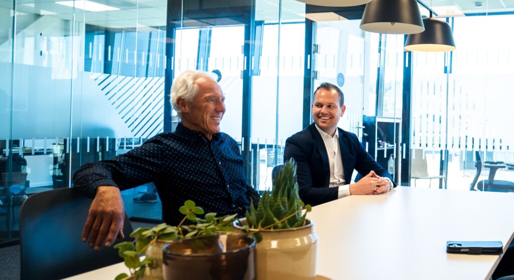Two men seated at a white conference table in a glass-walled office, smiling and chatting; a potted plant sits in the foreground.