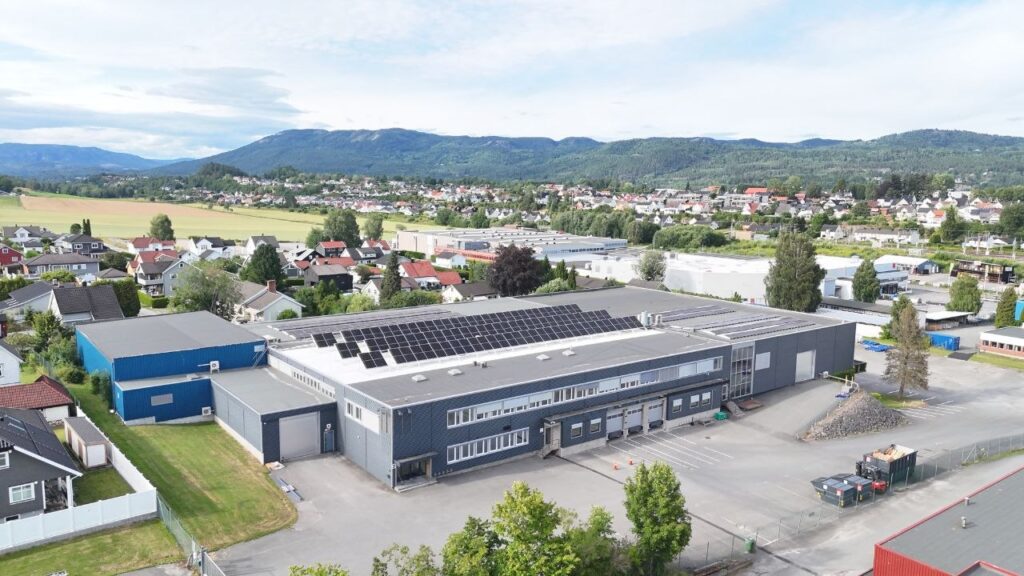 Industrial complex with solar panels on the roof in a small town, hills in the background.
